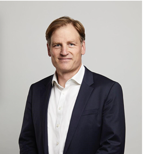 A man with short light brown hair wears a dark suit jacket over a white dress shirt. He stands against a plain light gray background, looking at the camera with a neutral expression, as if preparing to present in the VTE Webinar Series.