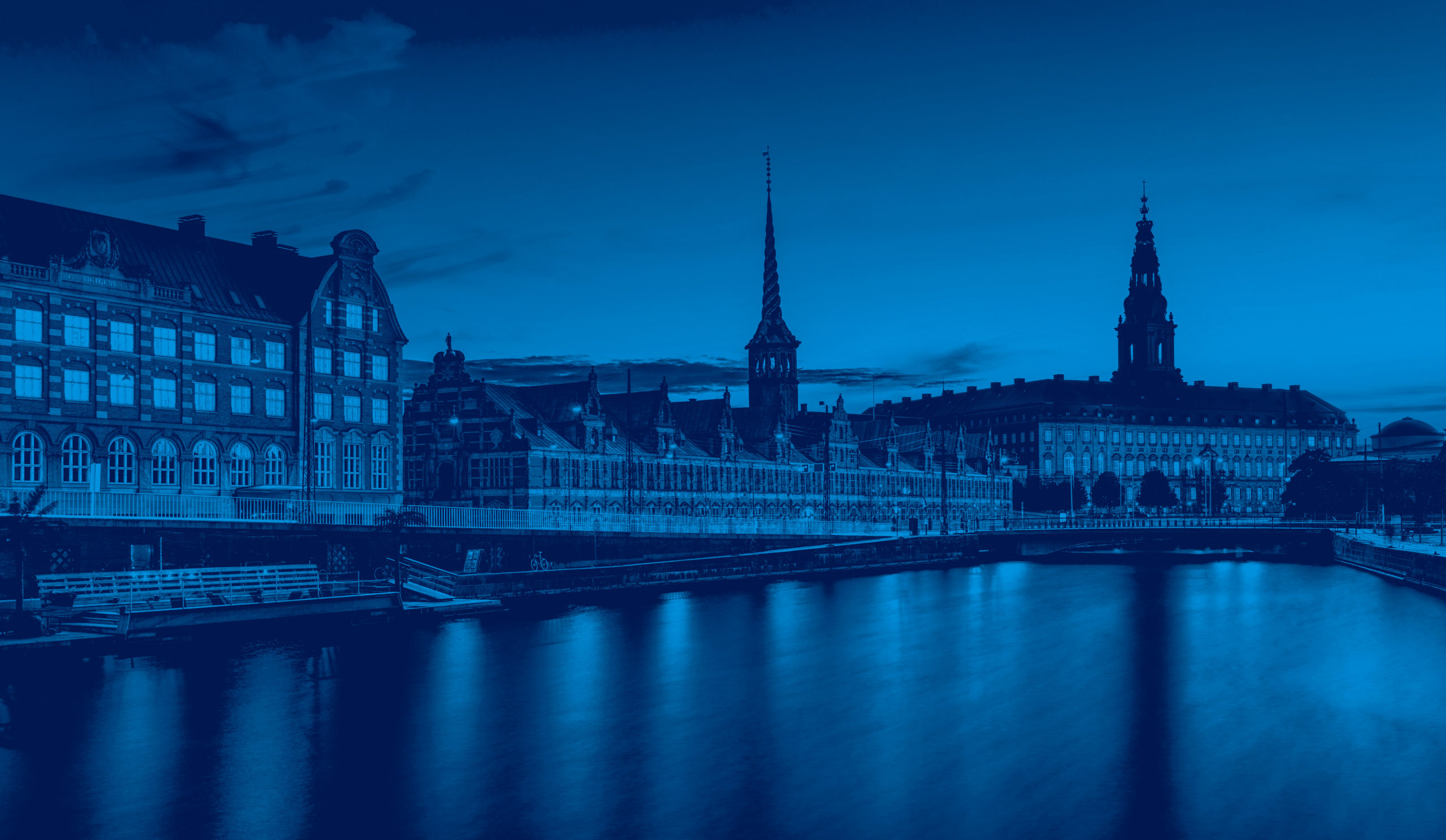 Cityscape with historic buildings and church spires reflected in a river under a cloudy sky. The image has a strong blue tint, giving it a monochromatic, nighttime appearance.