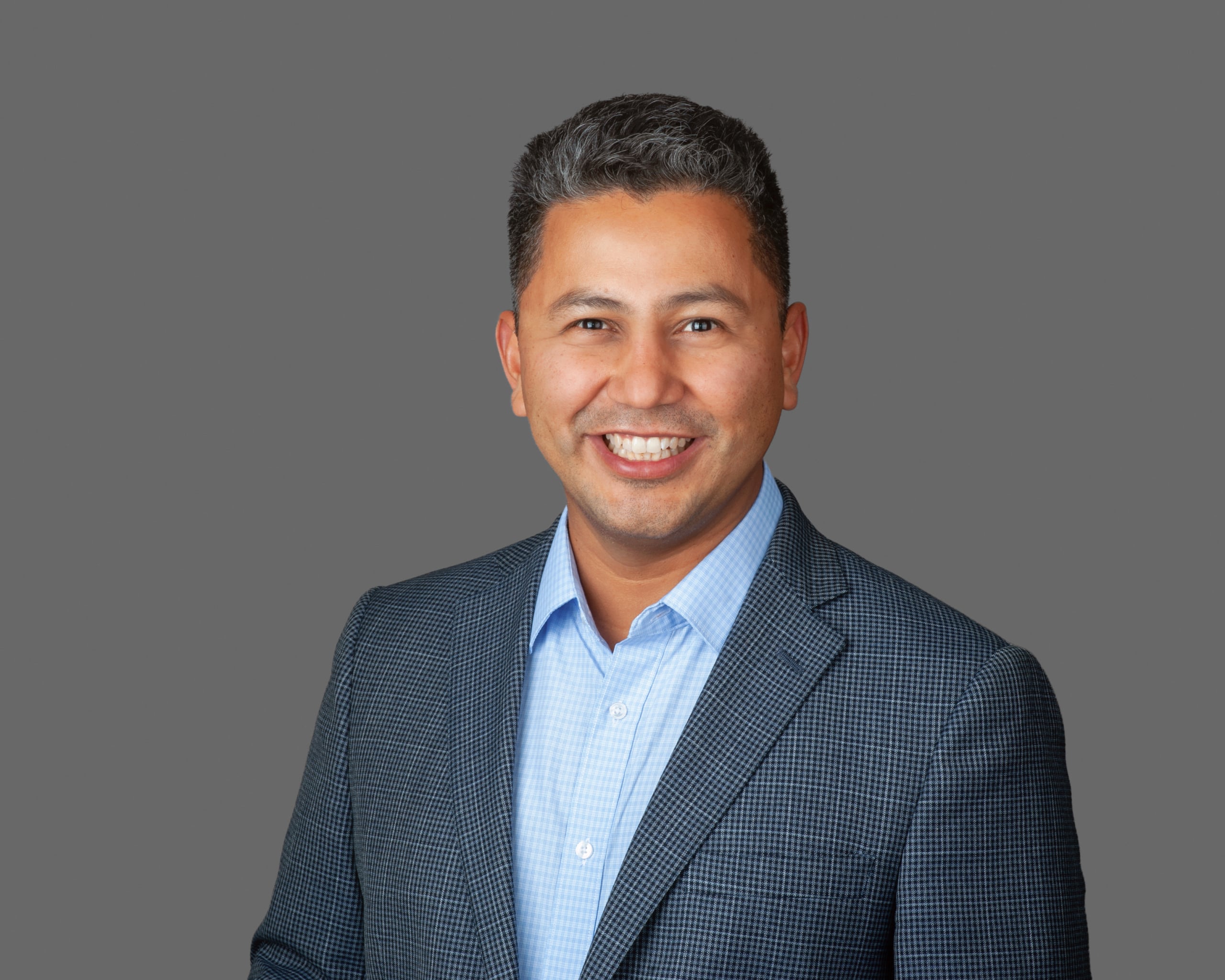 Miguel Miranda smiling while wearing a blue button-down shirt and a patterned blazer against a gray background.